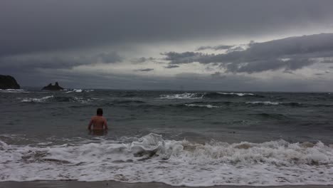 A-slow-motion-shot-of-a-beareded-man-walking-into-stormy-waves-and-swimming-in-winter