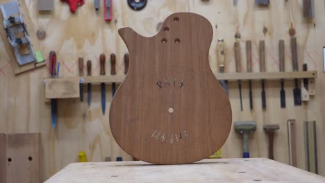 Unfinished-Custom-Built-Guitar-Body-With-Cut-Runes-Standing-on-Worktable-with-Woodworking-Tools-Hanging-on-the-Wall-in-Background-at-Luthier's-Workshop