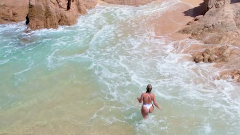 Shot-of-a-beautiful-woman-in-a-white-bikini-walking-along-the-sandy-and-brown-rocky-shore