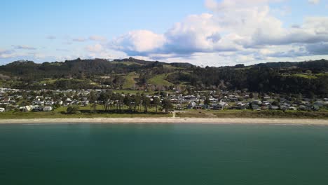 Flying-down-beach-side-town-of-Cooks-Beach,-New-Zealand-with-mountains-in-the-background