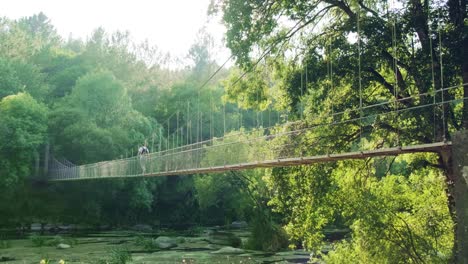 Establishing-shot-caucasian-man-crossing-rope-suspended-bridge,-Idyllic-forested-landscape