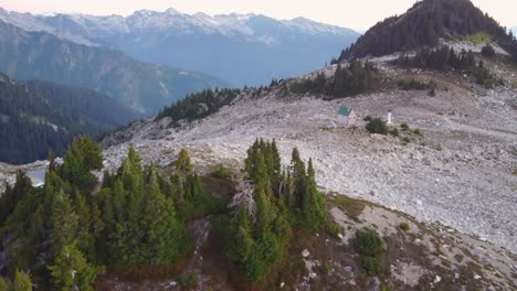 Slow-Tilting-Up-Aerial-Drone-Shot-of-Pine-Trees-and-Lone-Cabin-at-Sunset-on-Mount-Brew-with-Snow-Capped-Mountains-in-Canada-BC-4K