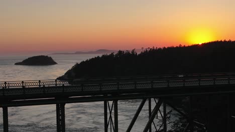 Slow-moving-aerial-shot-of-a-car-driving-across-Deception-Pass-State-Park-with-the-glow-of-the-sunset-in-the-backdrop