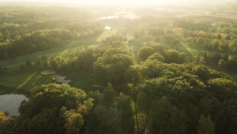 Aerial-View-of-Golf-Course-Field-at-Sunrise,-Green-Grassland-Lawn-Lands-and-Trees-in-Vast-Nature-Park-Forest-in-Beautiful-Sunny-Autumn-Day,-Sunlight-Flare-in-Horizon