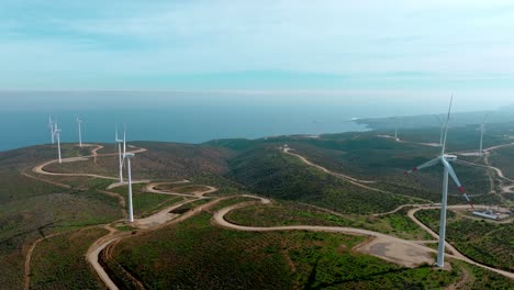 Drone-flying-towards-windmills-spinning-on-forested-hilltop,-wind-farm-in-northern-Chile-oceanfront