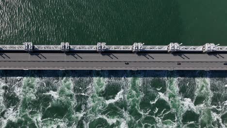 Stunning-wide-aerial-birdseye-view-of-a-storm-surge-barrier-with-a-road-and-passing-cars-with-calm-water-on-one-side-and-rough-foamy-water-on-the-other