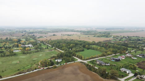 Aerial-High-Altitude-View-of-Rural-Countryside-Neighborhood-Area,-Crossroad-Surrounded-by-Local-Homes-Green-Golf-Course-Field-and-Vast-Agricultural-Lands,-Cloudy-Skyline-in-Horizon,-Pelham-Ontario