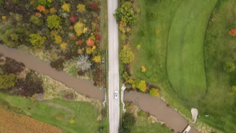 Aerial-Top-Down-Tracking-View-of-White-Car-Vehicle-Driving-Moving-along-Rural-Countryside-Road,-Route-Through-Nature-Autumn-Fall-Season-Landscape,-Green-Lands-Fields-and-Colorful-Trees-Around