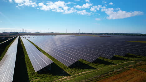 Aerial-Trucking-Shot-Of-Modern-Solar-Panel-Farm-Auf-Ländlichen-Feldern-Und-Windkraftanlagen-Im-Hintergrund-Im-Sommer