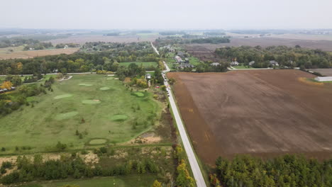 Aerial-View-of-Countryside-Nature-Landscape-at-Cloudy-Day-in-Autumn-Season,-Road-Through-Green-Golf-Course-Field-and-Vast-Agricultural-Lands,-Stationary-Vehicles-at-Roadside,-Skyline-in-Horizon