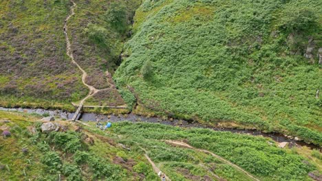 Aerial-video-footage-of-a-footpath-and-stream,-river-showing-a-small-bridge-structure-with-people-resting-along-the-riverbank