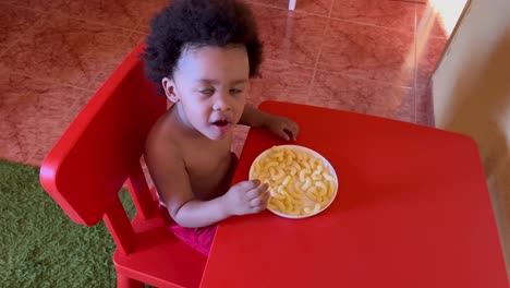Exotic-two-year-old-african-american-kid-eating-cheese-snack-seated-in-a-tiny-red-plastic-table