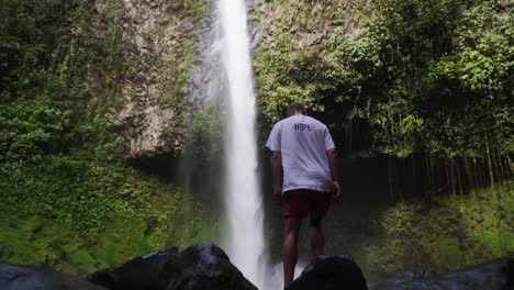 Shot-of-a-young-man-standing-and-lifting-his-arms-in-front-of-a-massive-waterfall-in-the-rain-forest-in-la-fortuna,-costa-rica