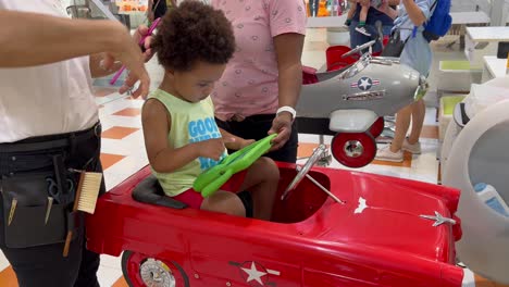 Lovely-two-year-old-child-in-the-hairdresser-seated-in-a-red-toy-car-focused-in-an-Ipad-next-to-his-mother,-wearing-a-sleeveless-shirt