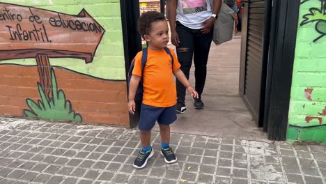 Adorable-two-year-old-african-european-child-livng-the-nuresy-after-his-fist-day-after-summer-vacations,-wearing-orange-shirt,-blue-shorts-and-a-backpack,-followed-by-mum
