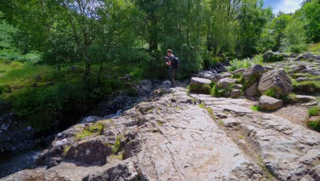 Young-boy-photographing-a-slow-moving-woodland-stream-with-ferns-on-the-riverbank-and-water-flowing-over-the-rocks