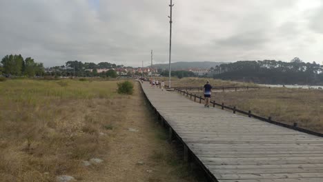 A-mature-adult-man-walks-behind-a-group-of-people-over-a-wooden-overpass-near-the-beach-one-cloudy-gray-morning,-panoramic-shot