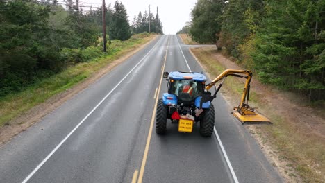 Tight-aerial-shot-following-a-tractor-using-a-flail-mower-to-level-the-overgrown-weeds-on-the-side-of-the-rural-road