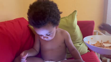 African-european-exotic-two-year-old-child-eating-nigerian-typical-food-by-himself-seated-in-the-sofa