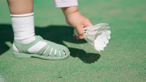 Toddler's-hand-picking-up-white-badminton-shuttlecock-from-the-ground-of-green-court-outdoors---close-up