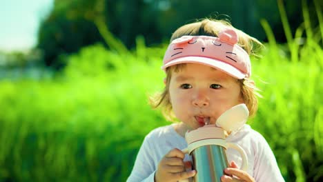 Adorable-american-child-girl-drinking-from-kids'-water-bottle-in-a-park-against-lush-green-bushes---slow-motion,-portrait