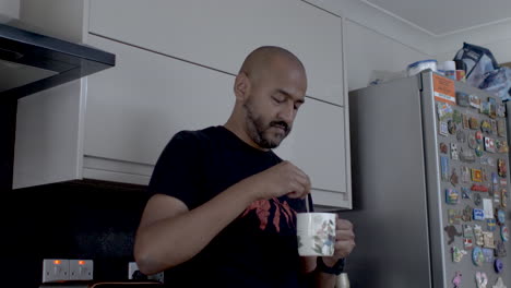 Bald-Adult-Male-Indian-Stirring-Tea-In-Mug,-Resting-Back-On-Kitchen-Counter