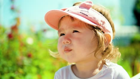 Dreaming-face-of-little-blonde-toddler-girl-wearing-pink-cap-and-looking-aside-in-a-garden-on-summer-sunny-day