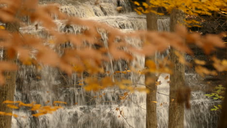 Rocky-waterfall-through-autumn-color-leaves-gently-swaying-in-the-wind,-Slow-motion