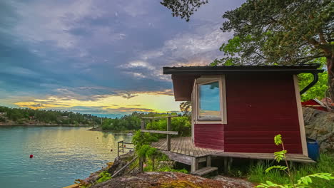 A-red-cottage-by-a-lake-in-Sweden-at-sunset-with-a-colorful-sunset-reflecting-off-the-water---motion-time-lapse