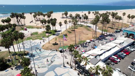 Old-Glory-flag-flying-over-the-boardwalk-and-skate-park-at-Venice-Beach,-Los-Angeles-California---orbiting-aerial-parallax-view
