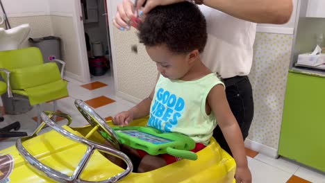 Exotic-two-year-old-african-europoean-child-in-the-hairdresser-for-his-first-hair-cut,-seated-in-a-yellow-toy-car