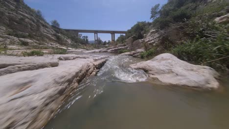 Low,-fast-FPV-drone-flight-down-narrow-rock-river-canyon-to-bridge