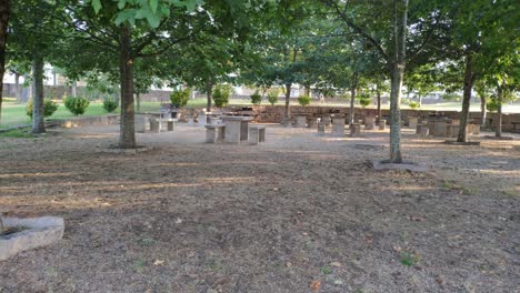 Granite-tables-in-a-picnic-area-with-barbecues-under-the-oak-trees-in-a-public-park-on-a-sunny-summer-afternoon,-rolling-shot-to-the-left