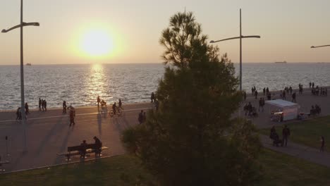Aerial---Low-shot-of-people-walking-on-Thessaloniki's-seaside-walkway-at-sunset