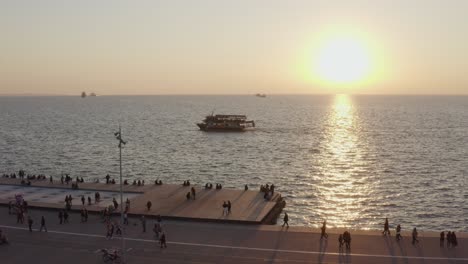 Aerial---People-walking-by-the-sea-in-Thessaloniki-with-a-boat-in-the-background-at-sunset