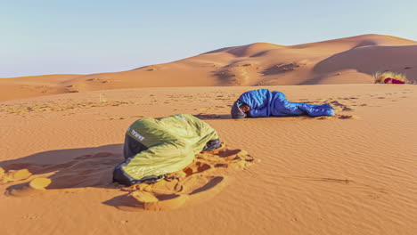 Two-people-camping-and-in-sleeping-bags-as-the-sun-rises-in-Morocco's-Sahara-Desert---time-lapse