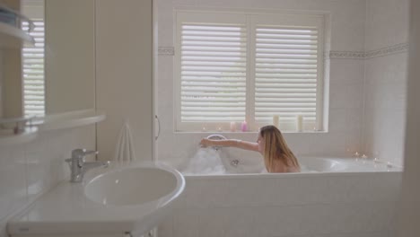 Wide-view-of-beautiful-woman-turning-of-faucet-and-laying-down-in-luxury-bathtub-covered-in-foam-which-is-surrounded-by-candles