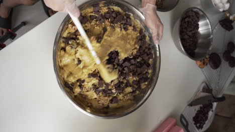 Overhead-Shot-Of-Cookie-Dough-With-Chocolate-Cubes-Being-Mixed-With-Spatula-In-A-Stainless-Mixing-Bowl