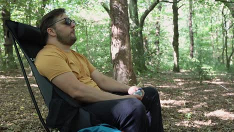 Man-with-sunglasses-eats-a-snack-and-rests-sitting-on-a-chair-at-a-campsite-in-the-forest