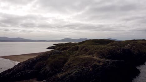 Aerial-low-rising-view-Ynys-Llanddwyn-island-Anglesey-coastal-walking-trail-with-Snowdonia-mountains-across-the-Irish-sea