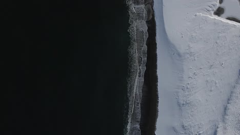Top-down-aerial-view-snow-covered-beach-with-black-sea-waves-on-coast