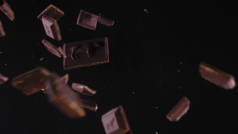 Dark-chocolate-block-falls-on-a-black-background,-explodes-and-parts-fly-in-the-air---extreme-slow-motion-top-shot