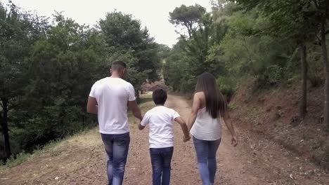 Mother-and-father-walking-on-dirt-road-in-the-nature,-holding-the-hands-of-their-child-son-in-the-middle,-all-dressed-in-white-and-blue
