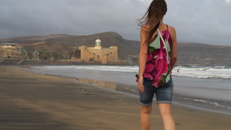 Cinematic-shot-at-dawn-of-a-woman-walking-on-the-beach-of-Las-Canteras-and-in-the-background-you-can-see-the-Alfredo-Kraus-Auditorium