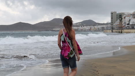 Cinematic-shot-at-sunrise-of-a-woman-walking-along-Las-Canteras-beach-and-seeing-the-houses-in-the-area