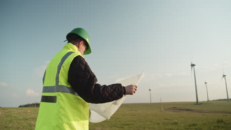 An-engineer-in-a-reflective-vest-stands-with-design-plans-at-a-wind-farm