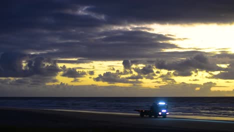 Dramatic-and-cinematic-blue-and-yellow-sunrise-sunset-over-a-calm-pacific-ocean-beach-scene-with-a-silhouetted-brightly-lit-flatbed-truck-driving-along-the-beach