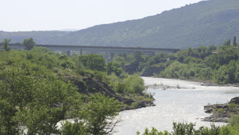 Coches-Circulando-Por-El-Puente-Que-Cruza-El-Río-A-La-Luz-Del-Día-Con-Vistas-A-Las-Montañas-Al-Fondo