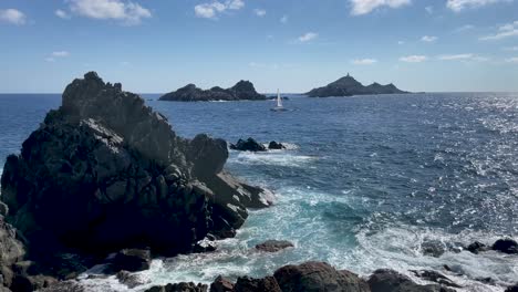 Sea-waves-crashing-in-to-rocky-coastline-in-corsica-island,-while-sail-boat-crossing-the-sea