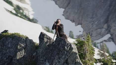 Photographer-pivots-with-camera-on-tripod-on-alpine-mountain-ridge,-Tim-Durkan-medium-tracking-helicopter-counterclockwise-orbit-SLOW-MOTION